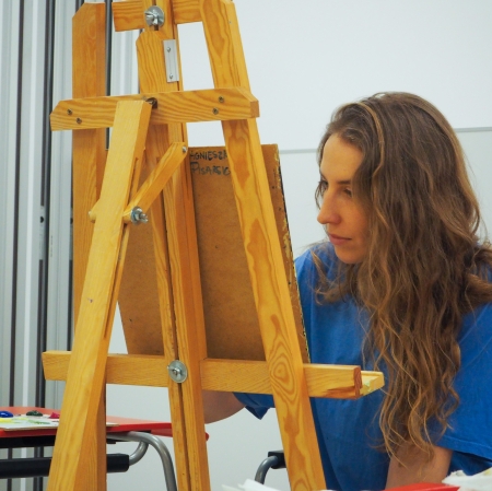 Osoba o długich włosach, siedząca przy sztaludze i malująca.