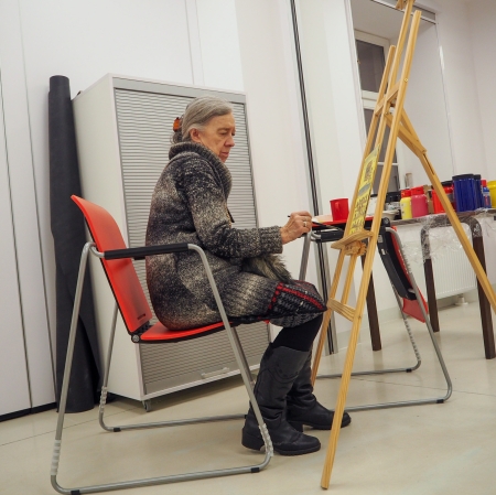 Starsza osoba siedząca na krześle przy sztaludze. Osoba nabiera farbę i zaczyna malować.