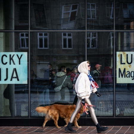 kobieta w białej kurtce z kapturem, z rudym psem przechodząca obok wystawy